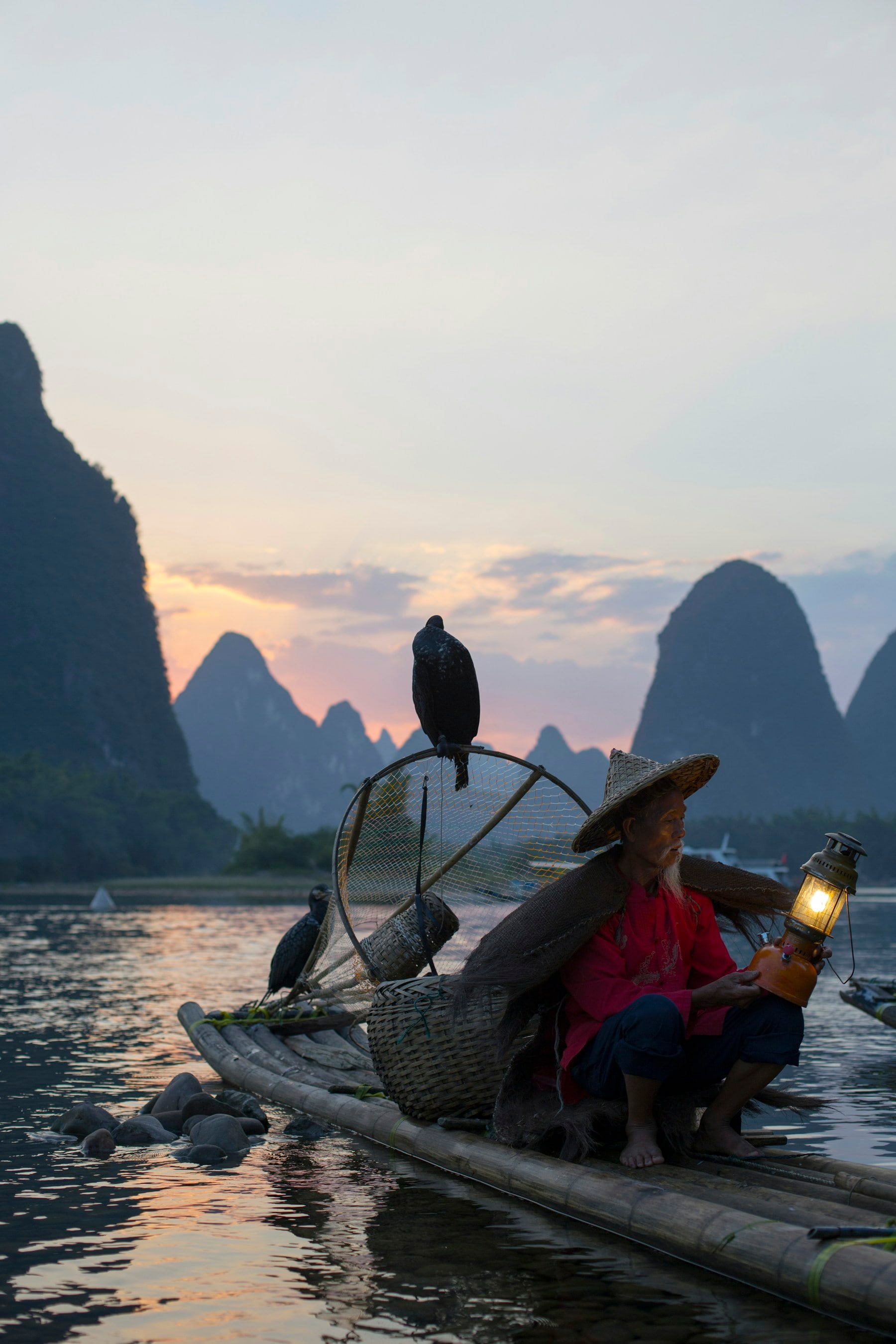 A cormorant fisherman sitting on a bamboo raft with a lantern at dusk near the Li River